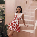 Woman wearing a red and white patchwork floral mini dress with side cutouts and wide straps, leaning against pastel stairs in a sunlit outdoor setting.